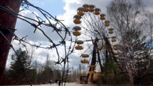 Arame farpado em primeiro plano foca uma roda-gigante abandonada e enferrujada. Uma roda-gigante amarela está parada entre árvores secas sob céu nublado.