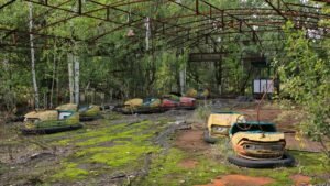 Carrinhos de bate-bate enferrujados e abandonados estão dispersos sobre um chão coberto de musgo e folhagem nua. O brinquedo está sob uma estrutura de metal enferrujada, cercado por árvores densas.