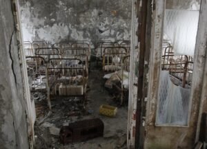 Quarto de creche abandonada repleto de berços de ferro enferrujados. As paredes apresentam tinta descascada e o chão está coberto de detritos, restos de colchões e brinquedos velhos. A cena é vista através de uma porta com vidros partidos.
