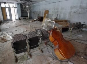 Um contrabaixo acústico de madeira encostado em poltronas estofadas antigas no centro de uma sala abandonada. Sobre uma das poltronas há o retrato em preto e branco de um cachorro. O chão e as paredes estão deteriorados, com entulho, móveis quebrados e cabides espalhados pelo ambiente.