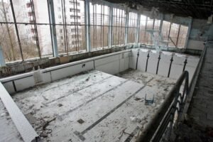 Piscina olímpica coberta inteiramente vazia e abandonada, com o fundo repleto de sujeira e escombros. Ao fundo, vê-se a plataforma de saltos e grandes janelões que mostram edifícios residenciais ao ar livre.
