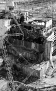 Foto em preto e branco do Reator 4 de Chernobyl após a explosão. Mostra a fachada severamente danificada da usina, com escombros acumulados na base e estruturas metálicas retorcidas.