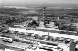 Vista aérea panorâmica em preto e branco do complexo da Usina Nuclear de Chernobyl após a explosão. No centro do extenso complexo industrial, destaca-se a secção destruída do Reator 4, com danos visíveis na estrutura ao lado da alta chaminé de ventilação.