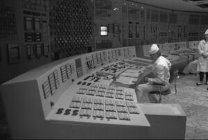 Foto em preto e branco da sala de controle do Reator 4 antes do acidente. Um operador de uniforme e chapéu brancos está sentado diante de um painel curvo repleto de botões e mostradores. Ao fundo, as paredes estão compostas por painéis de controle e outros trabalhadores observam o ambiente.